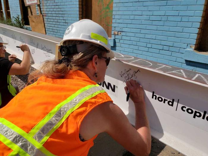 A woman in a hardhat signs her name on a steel beam.