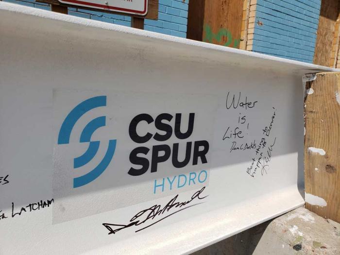 A steel beam, painted white, with the logo of the CSU Spur Hydro building and signatures.