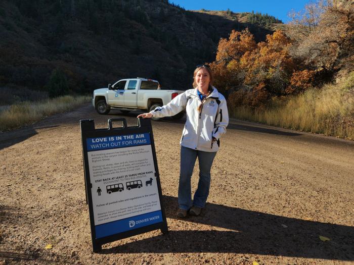 A woman in a jacket with the Denver Water logo stands next to a sign warning people to keep their distance from wildlife in Waterton Canyon. A Denver Water truck is in the background. 