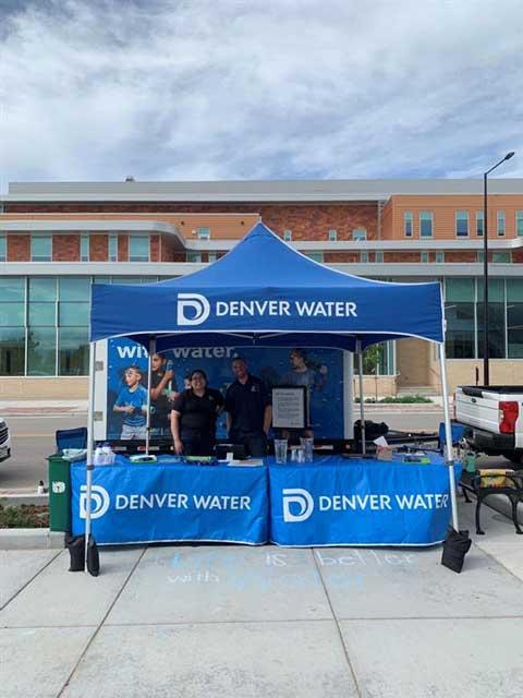Denver Water set up at a community event with a blue tent and blue tablecloths with logos. Smiling employees ready to talk with the public.