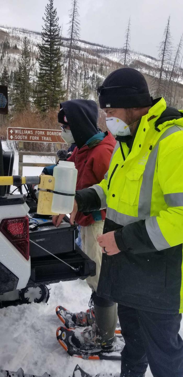 Two men stand next to a pickup full of testing equipment. It's cold and snowy. They're wearing masks and heavy coats.