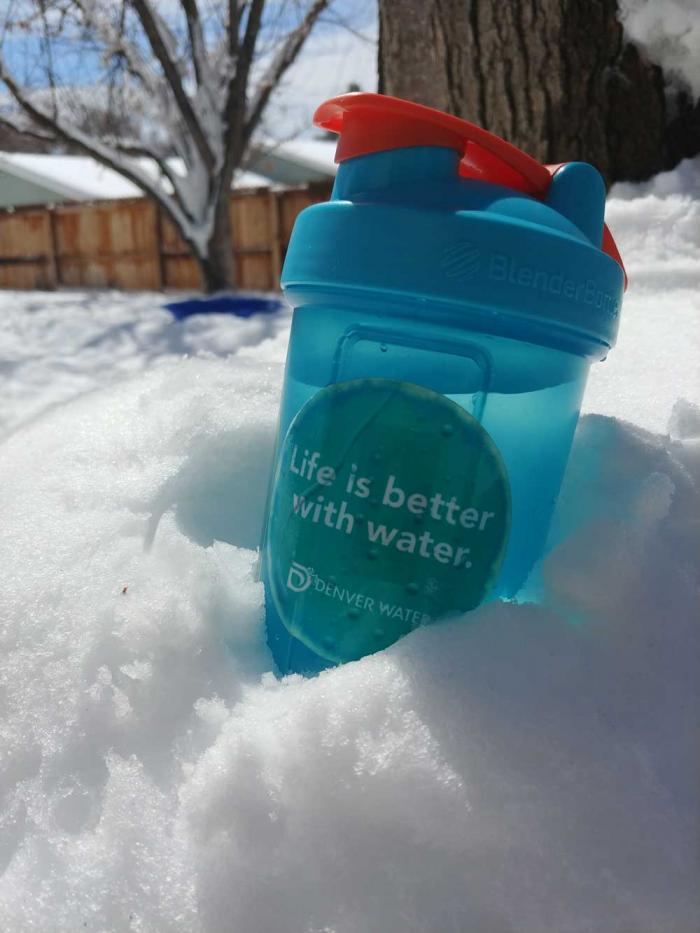 A water bottle, with a sticker that says "Life is Better With Water" and a Denver Water logo, rests in a pile of snow.