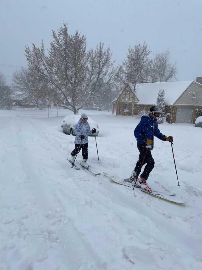 Two people cross-country ski down a street amid the snow. 