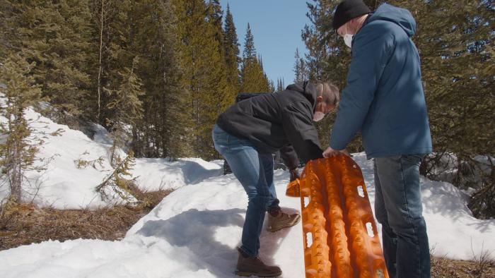 Two men lower an orange pad into the snow to form a bridge over a drainage for the utility terrain vehicle to drive over.