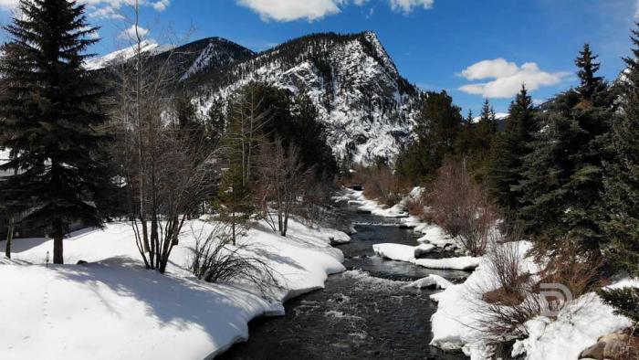A dark creek flows through snow-covered mountains with trees under sunny, blue skies.
