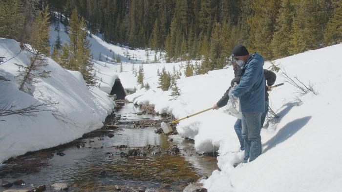 Two men stand in the snow next to a mountain creek. One holds a pole, attached to a bottle that will scoop up a water sample.