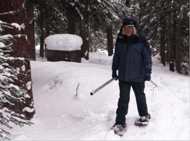 A man holding a metal tube snowshoes through the forest. 