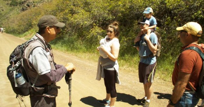 A man in a brown uniform shirt, backpack and hiking stick talks with a family on a trail.