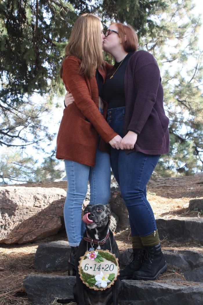 Kait Counter (left) and Casey celebrate their engagement with their beloved pup, Lola, who sports a sign announcing the couple’s original 2020 wedding date. Photo credit: Kait Counter.