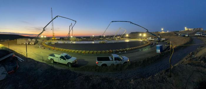 A work site lit by construction lights, with trucks and concrete pumps, and the dawn in the distance.