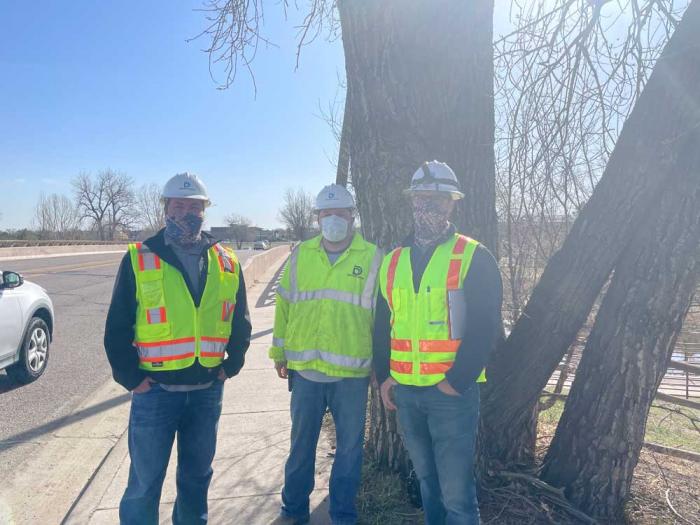 Three men in Denver Water hard hats and safety vest stand in front of a tree by the side of a road.