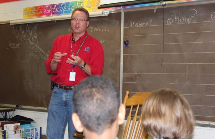 A man in a red shirt with a Denver Water logo stands in front of a classroom chalkboard talking to children.