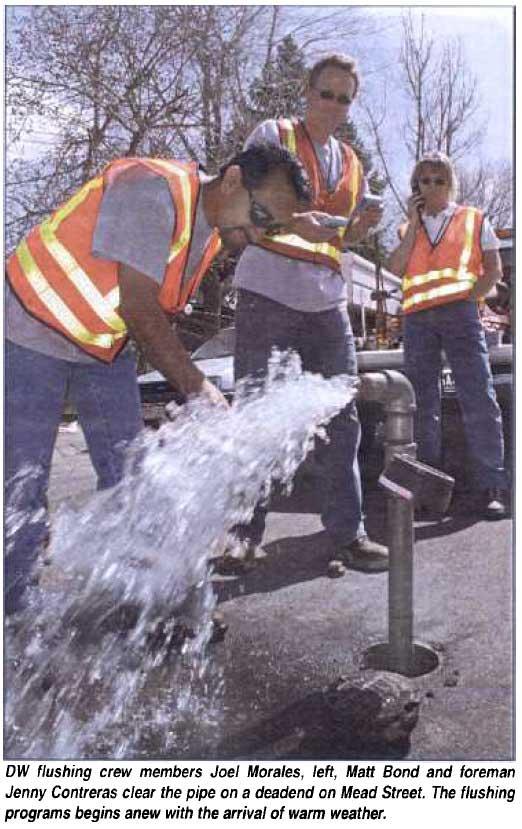 Three men in safety vests monitor water flushing from a hydrant.