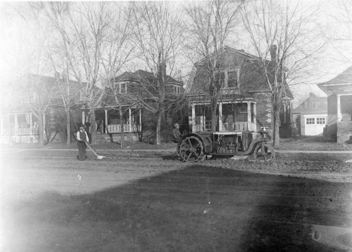 Two men guiding a tractor down a dirt street, with old brick houses in the background. 
