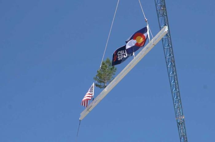 A white steel beam with flags fluttering in the air dangles on cables below a crane.