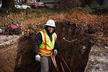 A worker stands in a hole, smiling, holding a coil of copper pipe.