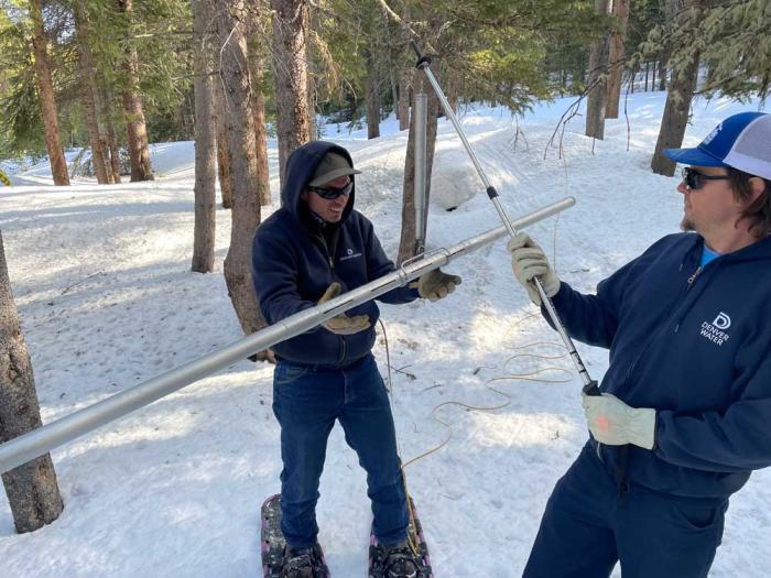 Three men in the woods filled with snow, one holds a hollow tube filled with snow while another squints at the measurement on the side.
