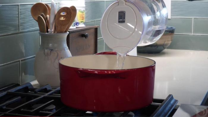 Filtered water is poured from a pitcher into a big red pot on a stove in a kitchen.