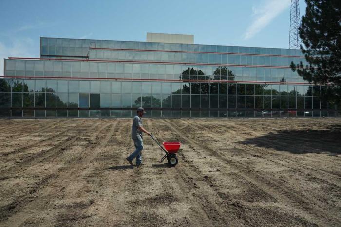 A man walking a seeder through a dirt field with an office building in the background. 
