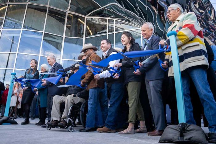 People lined up in front of a building holding large scissors, ready to cut the blue ribbon strung in front of them. 