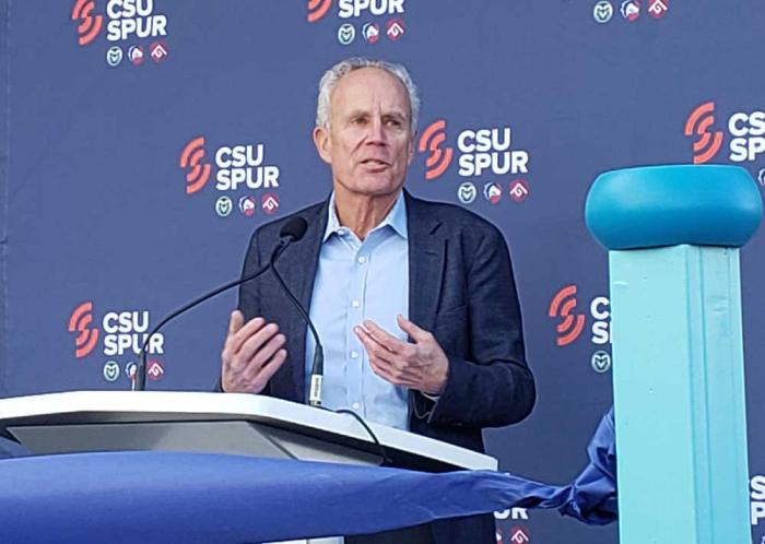 A man speaks at a podium, behind him is a drap saying "CSU Spur" with logos. 