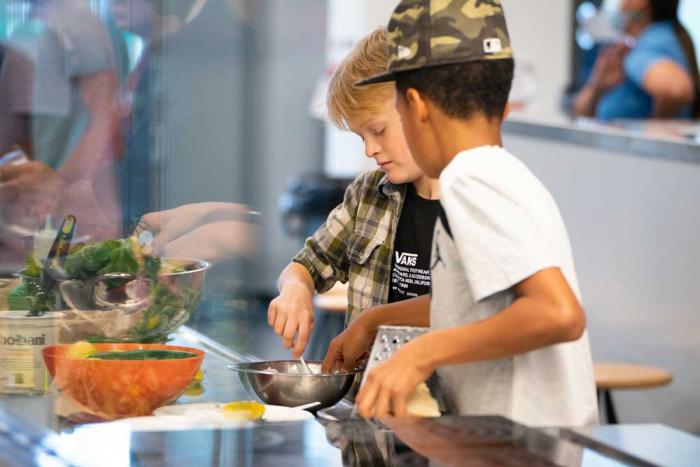 Two boys in a kitchen, peering into a pot. 