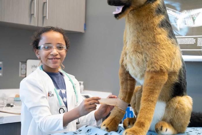 A girl in a white lab coat wraps a bandage around the leg of a stuffed dog. 