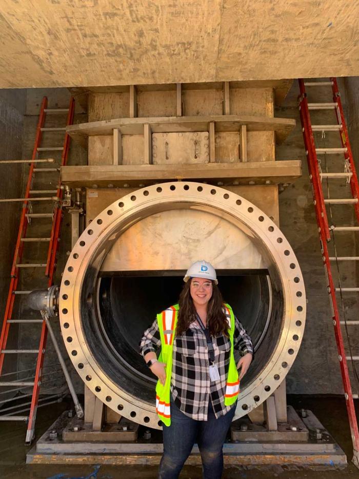 Woman in a Denver Water hard hat standing in front of a valve about 6 feet in diameter embedded in a wall. 
