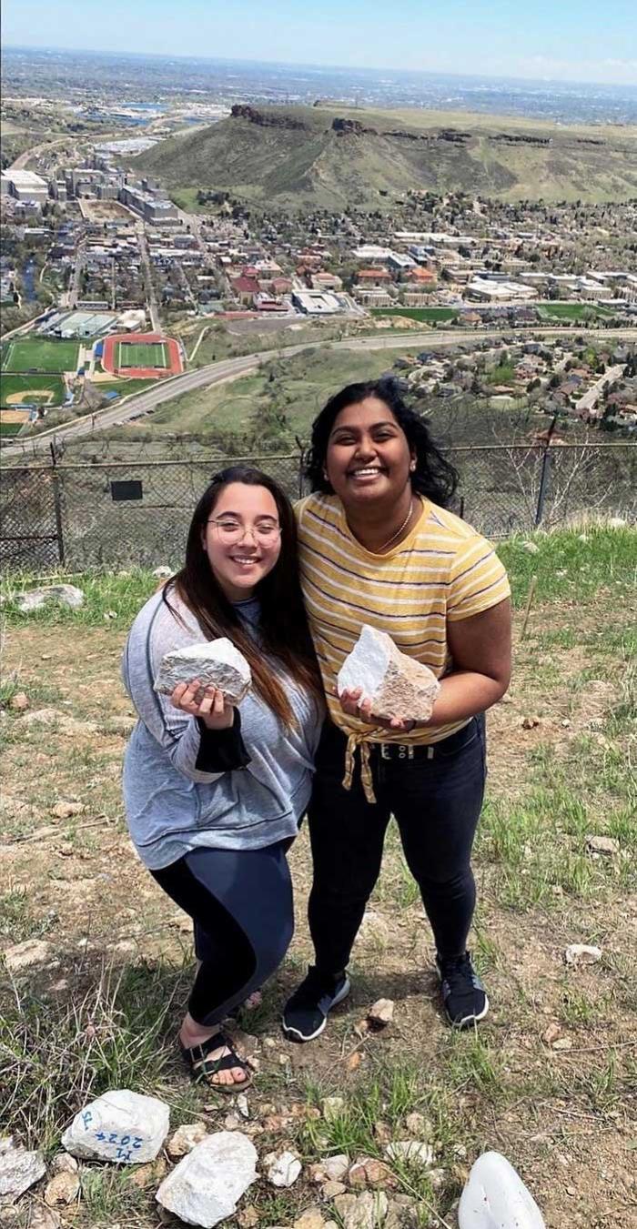 Two women on a hillside, with the city in the background, holding white-painted rocks and smiling at the camera 