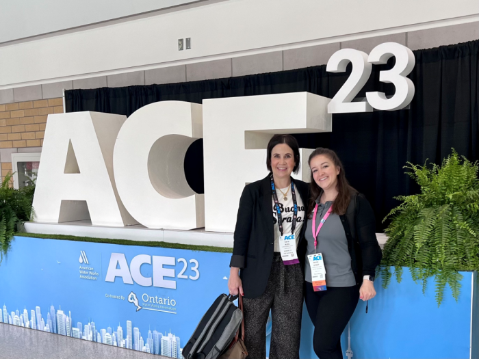 Two women stand in front of a sign that reads "ACE 23." They are wearing conference badges and are smiling at the camera. 
