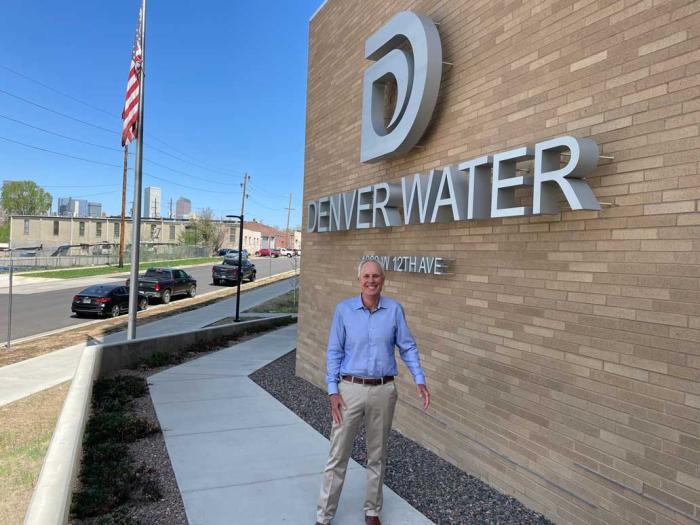 A man in a blue shirt and khaki pants stands in front of a building with a big D logo and the words "Denver WAter." 