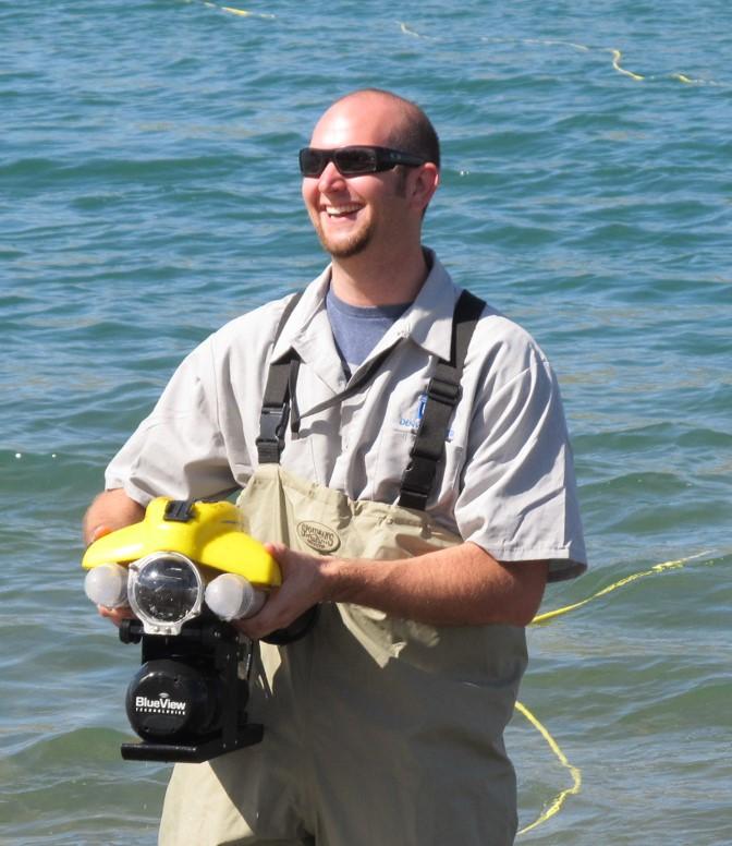 A man wearing sunglasses and smiling, the background is water and he's holding diving gear.