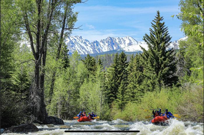 Rafters on a white-water river with trees, snow-capped mountains and blue sky. 