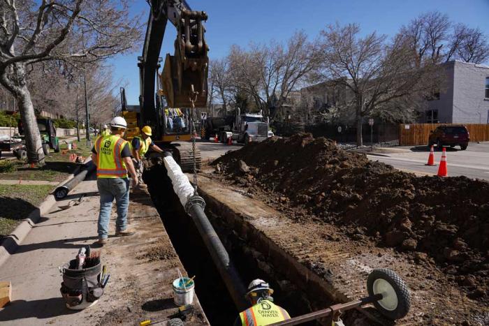A group of men in Denver Water safety vests and hard hats carefully lower a pipe into a trench in the street, assisted by a crane.