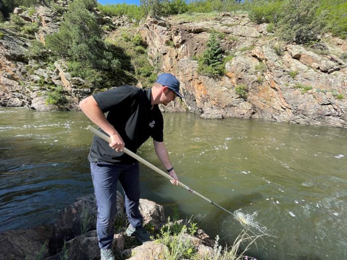 A man stands on a stream bank with a long pole in his hands. At the end of the pole is a cup, being dipped into the water to collect a water sample. The man is wearing a shirt and hat with the Denver Water logo.