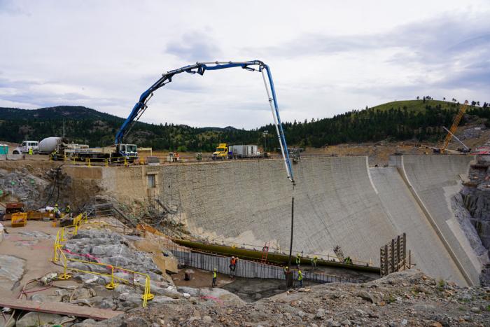 The side of a large grey dam takes up most of the picture. In the foreground is a pumper truck, the hose arching above the top of the dam and pouring concrete into a base where workers guide it into place. 