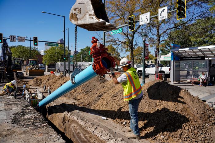 Workers carefully guide a blue pipe into a trench on Colfax Avenue.