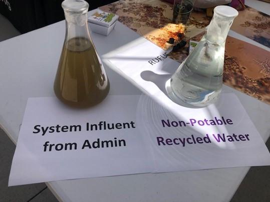 Two beakers sit side by side, one with brown water is labeled "system influent" the other with clear water is labeled "nonpotable recycled water." 