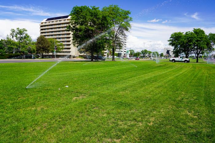 A huge green lawn with sprinklers spraying water with large office buildings in the background. No one plays on this lawn.
