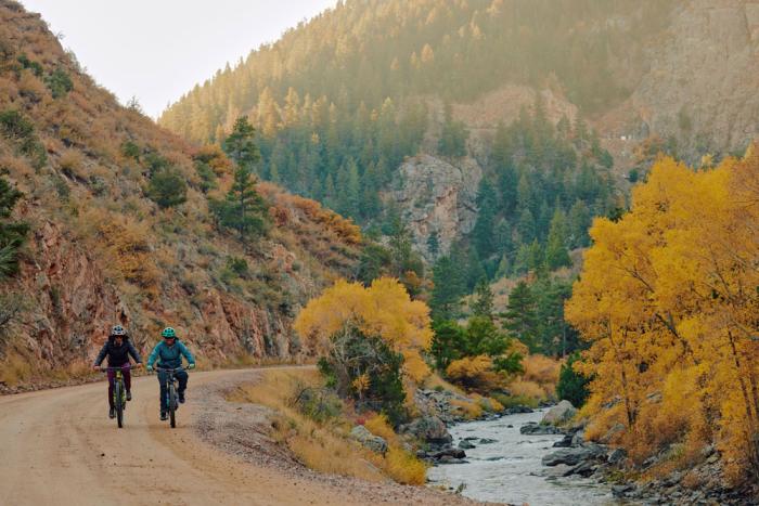 Two people ride bikes on a dirt road, next to a stream in a canyon. It's fall and the trees are turning yellow. 