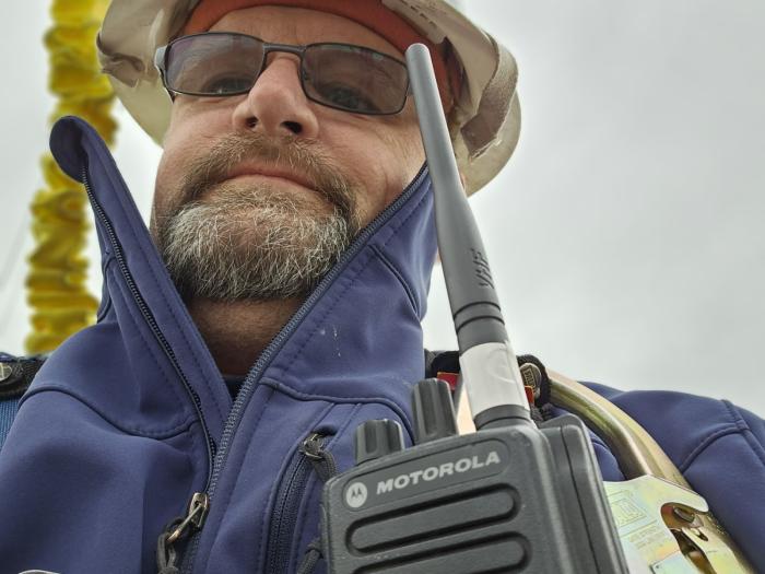 A man wearing a safety harness, hard hat, glasses and a radio looks into the camera. 
