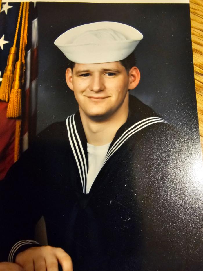 A young man in a U.S. Navy uniform, with a flag over his left shoulder. 