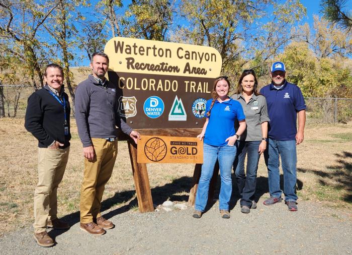 Five people line up next to a sign that says "Waterton Canyon Recreation Area" holding a wooden plaque carved with the words "Gold Standard Site" and "Leave No Trace" 