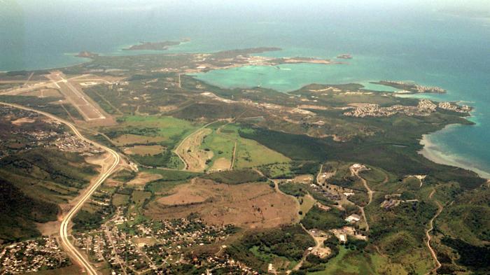 An aerial photo with beaches and blue ocean water seen at the top, and an airstrip and buildings seen at the bottom. 