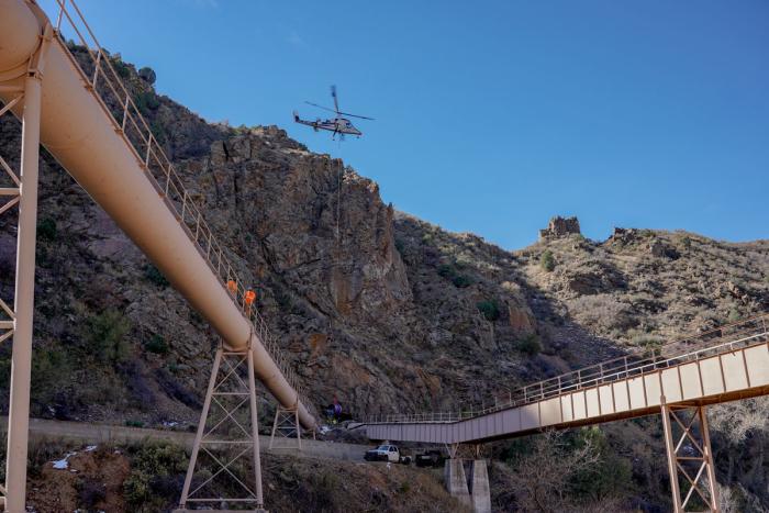 A helicopter hovers over a cliff, lowering a section of pipe down the side of the cliff. 