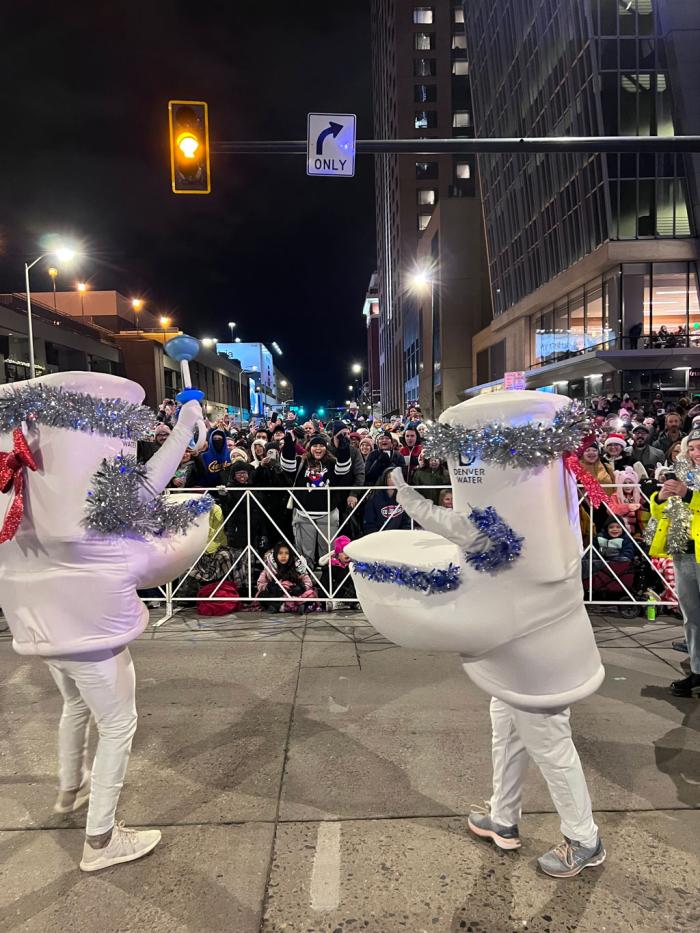 Two people in toilet costumes festooned with shiny Christmas garland dance in the street as part of a parade. 