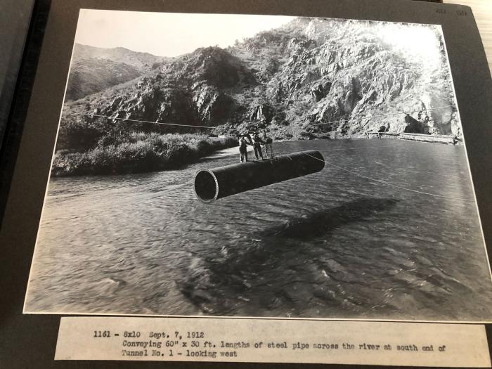 A historical, black and white image of a section of large pipe being pulled over a river on cable. 