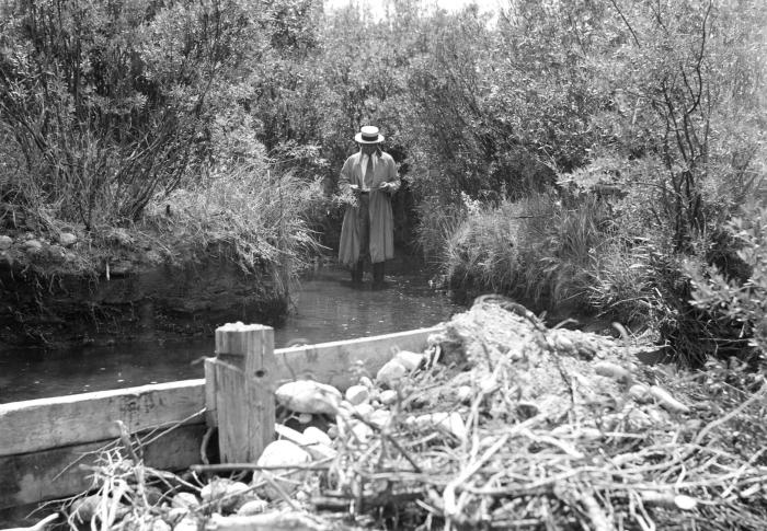 An old, black and white photo of a man in a trench coat and fedora, he's looking down at something in his hands and standing in water that's up on the rubber boots on his ankles. 