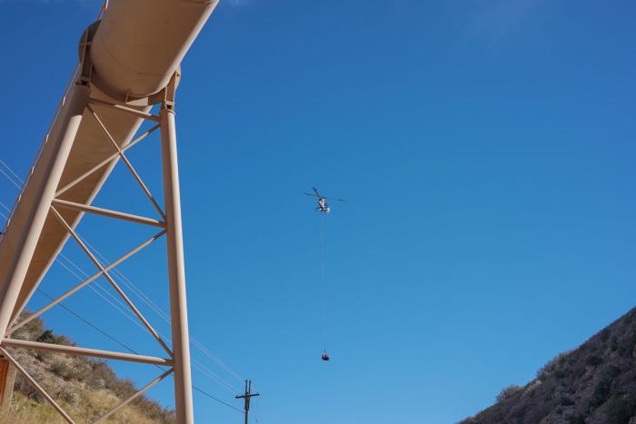 A helicopter with a section of pipe dangling underneath, flies through blue skies. 