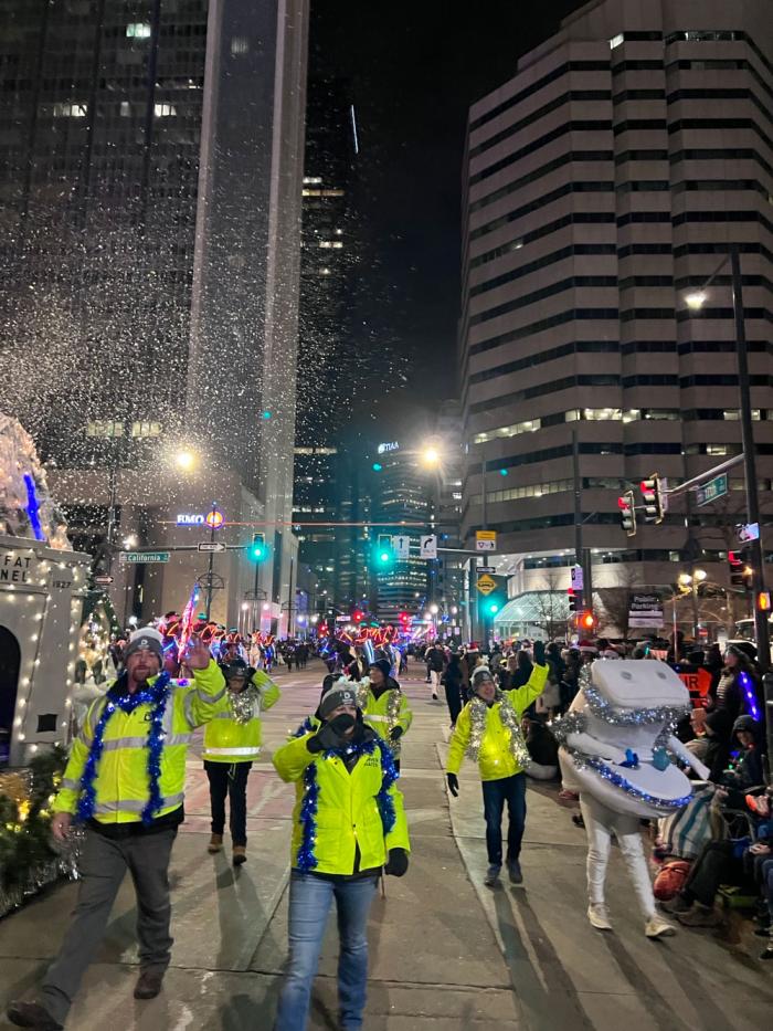 A group of people walking down the street parade, wearing yellow safety jackets, shiny Christmas garland, and waiving to the crowd. 